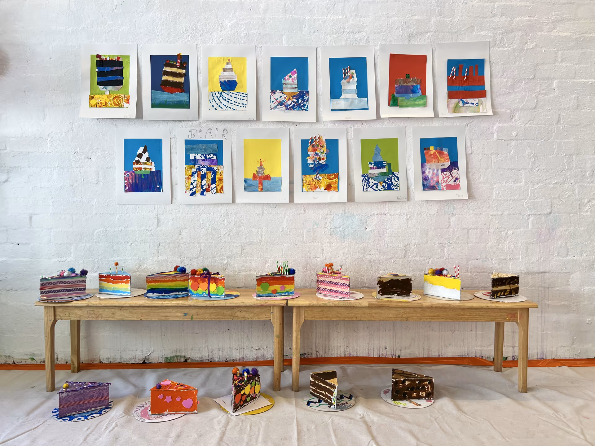 A wall of children's collage cake artworks above a long bench of 3D paper-and-paint cake sculptures, made during a Smudge Artspace holiday program